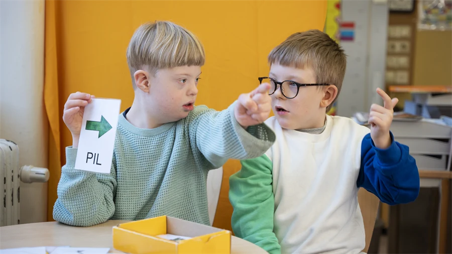 Bilden visar två barn som pratar och pekar. Ett av barnen håller i en lapp med en pil som pekar i samma riktning som barnen pekar.
