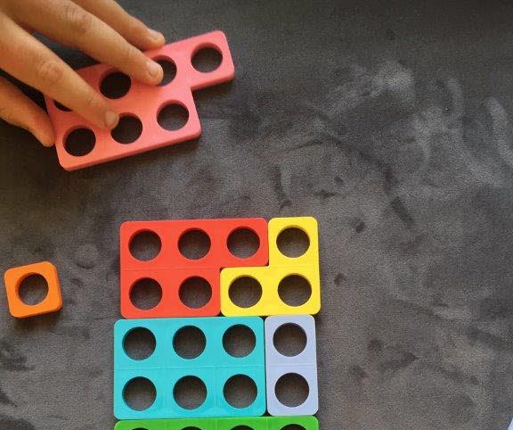En hand som håller i en plastskiva med sju hål i två rader. Nedanför syns andra plastskivor i olika färger som fogats samman så att de får jämna rader.