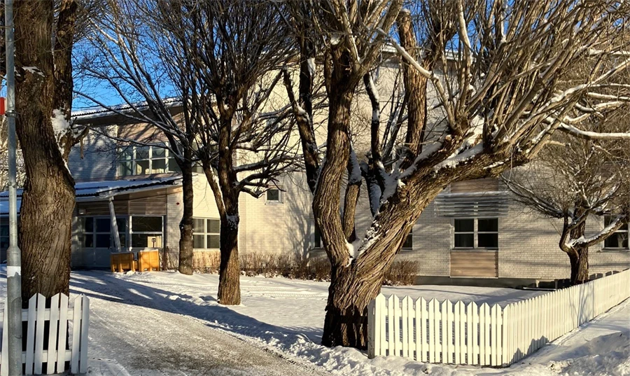 Snötäckt gångväg leder fram till en ljus, tvåvåningsbyggnad med stora fönster och fasad i ljus tegel- eller panelstruktur. Byggnaden omges av höga, bladlösa träd och ett vitt trästaket, i klart vinterljus.