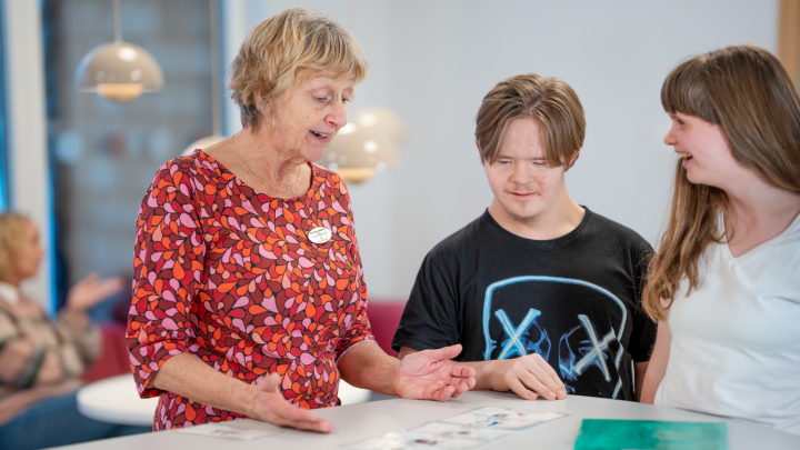 Bilden visar en vuxen person som pratar med två elever. De tittar tillsammans på en lapp med bildstöd.