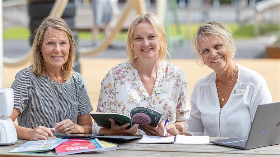 Tre kvinnor sitter vid ett bord utomhus. De tittar på varandra och pratar. På borden finns böcker, en dator och en liten robot. 