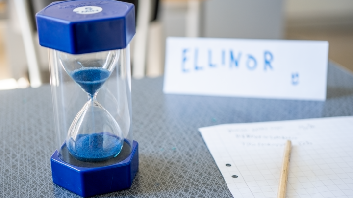 Ett timglas som står på ett bord, bredvid papper och en penna. I bakgrunden står en namnskylt