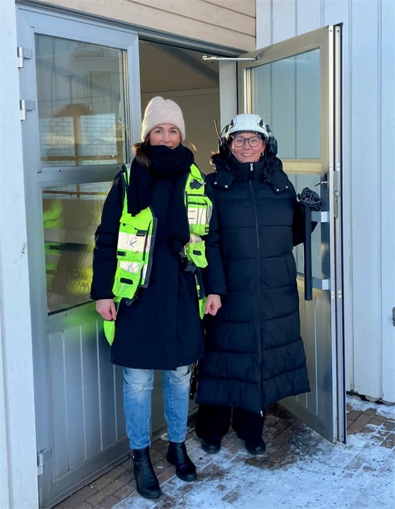 Två personer står vid en öppen ytterdörr till en ljus träbyggnad vintertid. Den ena bär mörk kappa, den andra mörk jacka med reflexväst och mössa. 