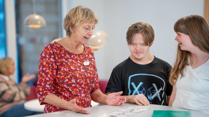 Bilden visar en vuxen person som pratar med två elever. De tittar tillsammans på en lapp med bildstöd.