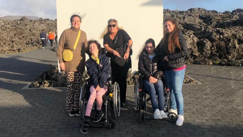 Två flickor och tre kvinnor står uppställda. Flickorna sitter i rullstol. Alla ler och tittar in i kameran. I bakgrunden syns ett stenigt lavalandskap.