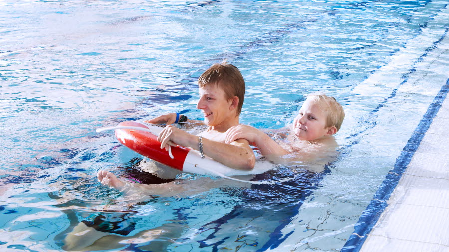 Två elever i en pool i en simhall. Den ene har en livboj runt sig, medan den andre håller i honom. I bakgrunden syns en elev med en annan livboj bredvid poolen.