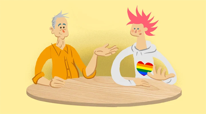 Två personer sitter vid ett bord och pratar. Den ene har grått hår och orange skjorta, den andre rosa hår och en tröja med ett regnbågshjärta på
