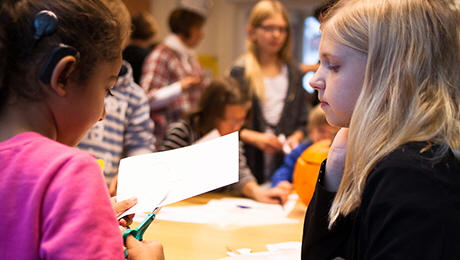 Två tjejer som sitter och kliipper i papper och fler personer i bakgrunden