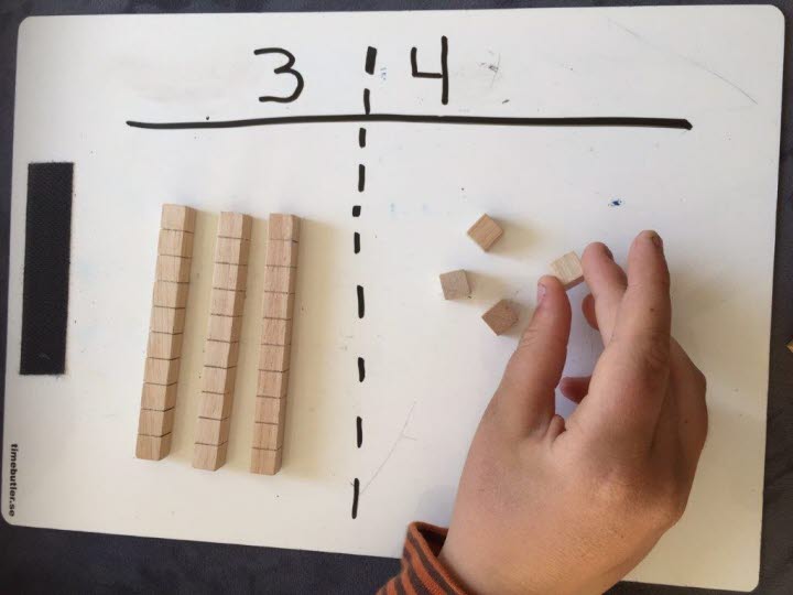 En hand som håller i en liten träkub som symboliserar 1. På ett papper finns en kolumn för tiotal med siffran tre och tre rader med tio träkuber i varje, samt en kolumn med siffran 4 och fyra stycken kuber
