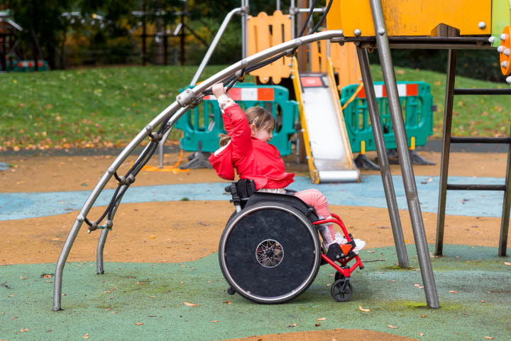 Fotografi som visar en flicka i rullstol som leker i en lekpark med hårt underlag.
