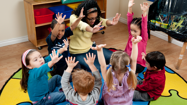 Barnen sitter samlade i en cirkel på golvet tillsammans med sin lärare. Alla håller upp sina händer i luften.