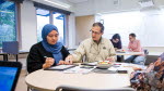 Elevcoach, Ghassan Alankar sitter vid ett bord med Nuria Rezae. De tittar ner på ett papper. I bakgrunden sitter elevcoach Kenth Edkvist och hjälper elev Can Polat. 