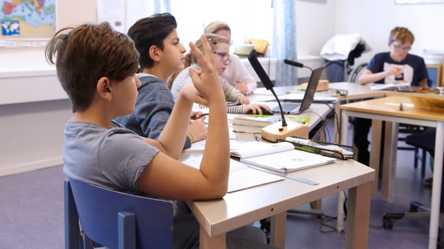 Bilden visar ett klassrum med elever där en elev räcker upp handen.