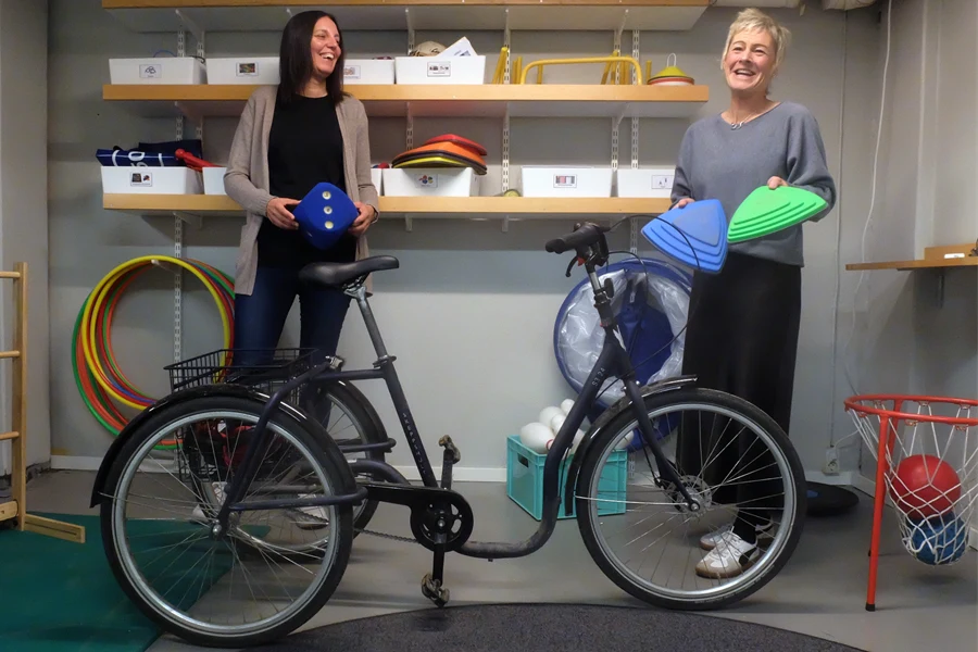 Två kvinnor visar upp en stor tärning, och en blå och en grön triangelformad sak. Framför dem står en trehjulig stor cykel. Bakom dem skymtar en hylla med lådor fyllda med olika saker.