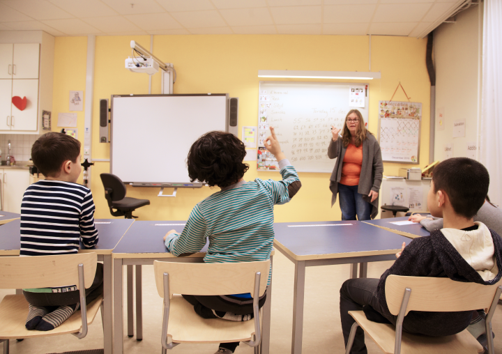 Tre elever i ett klassrum. En lärare står vid en whiteboardtavla.