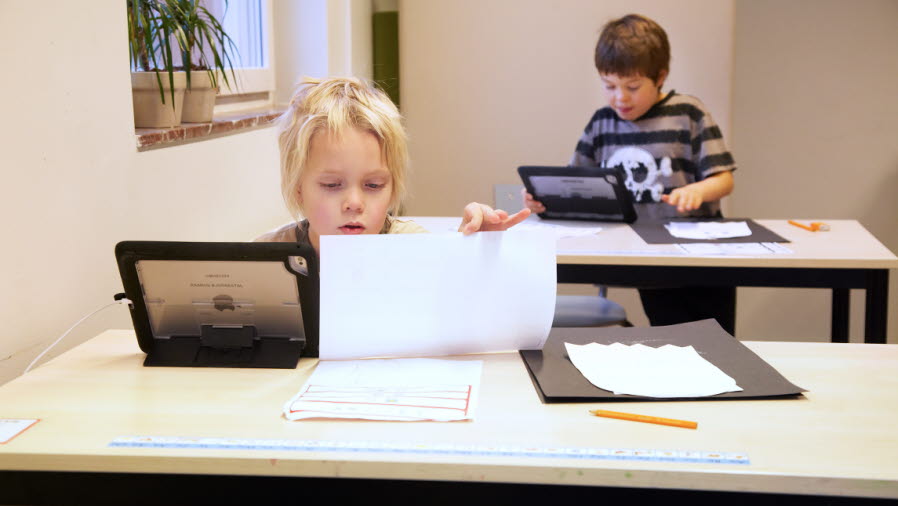 Två elever sitter vid var sitt skrivbord och arbetar. De har varsin surfplatta som komplement till papper och böcker.