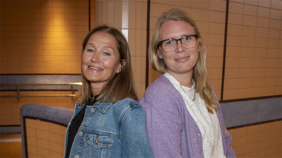 Två kvinnor står med ryggarna mot varandra. Båda ler och tittar in i kameran.