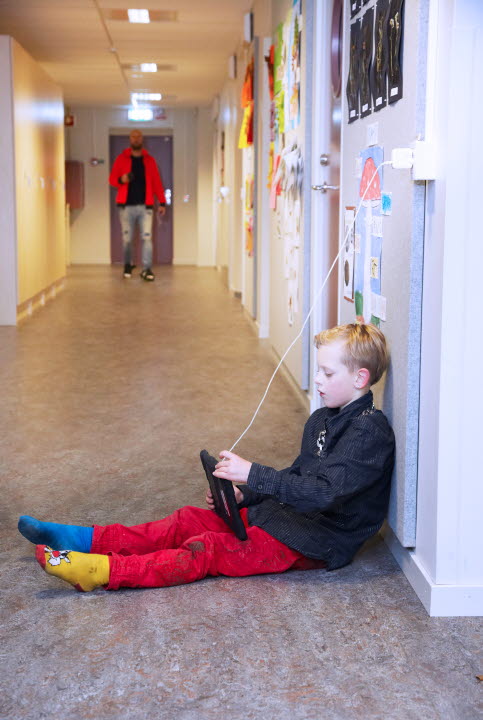 Bild på en pojke som sitter i en korridor på golvet med en surfplatta. Långt bort i korridoren kommer en vuxen.
